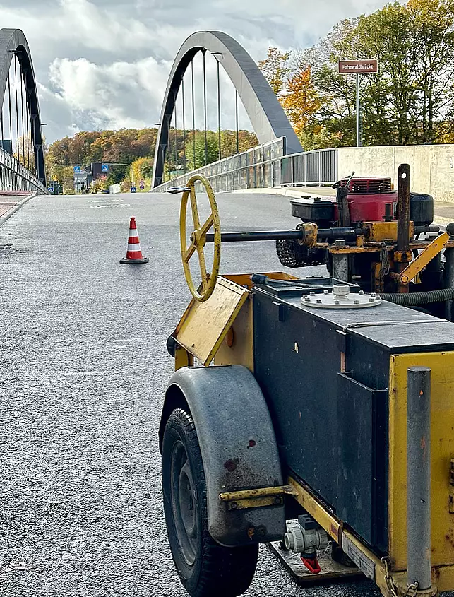 Prüfgerät auf einer asphaltierten Brückenfahrbahn mit Absperrkegel und bestehendem Straßenbauwerk im Hintergrund.